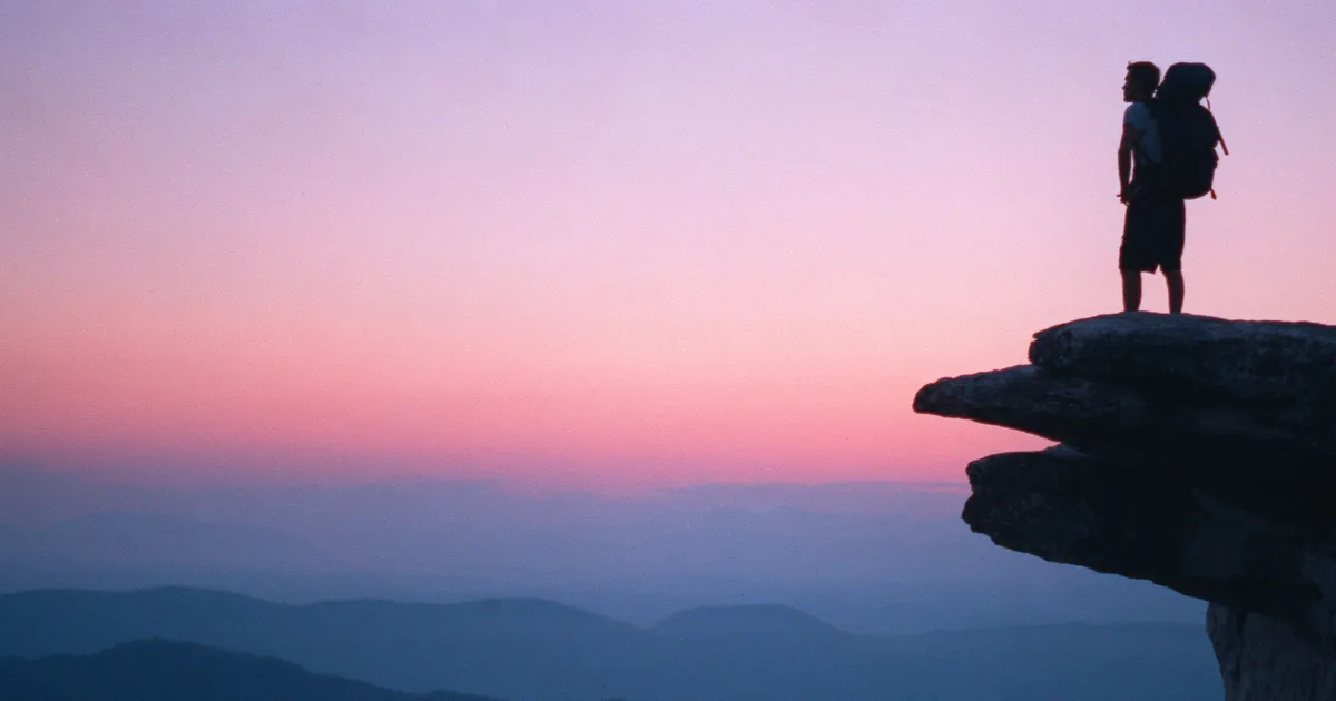 Silhouette of a man with backpack standing on McAfee Knob at sunset with mountains in the distance.