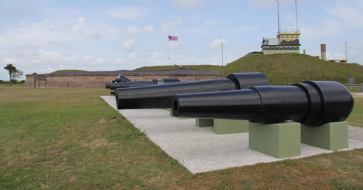 A row of cannon with Fort Moultrie in the background. A US flag is flying in the fort.