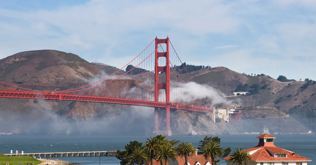 White buildings with red roofs at Crissy Field with blue bay and Golden Gate Bridge and fog behind.