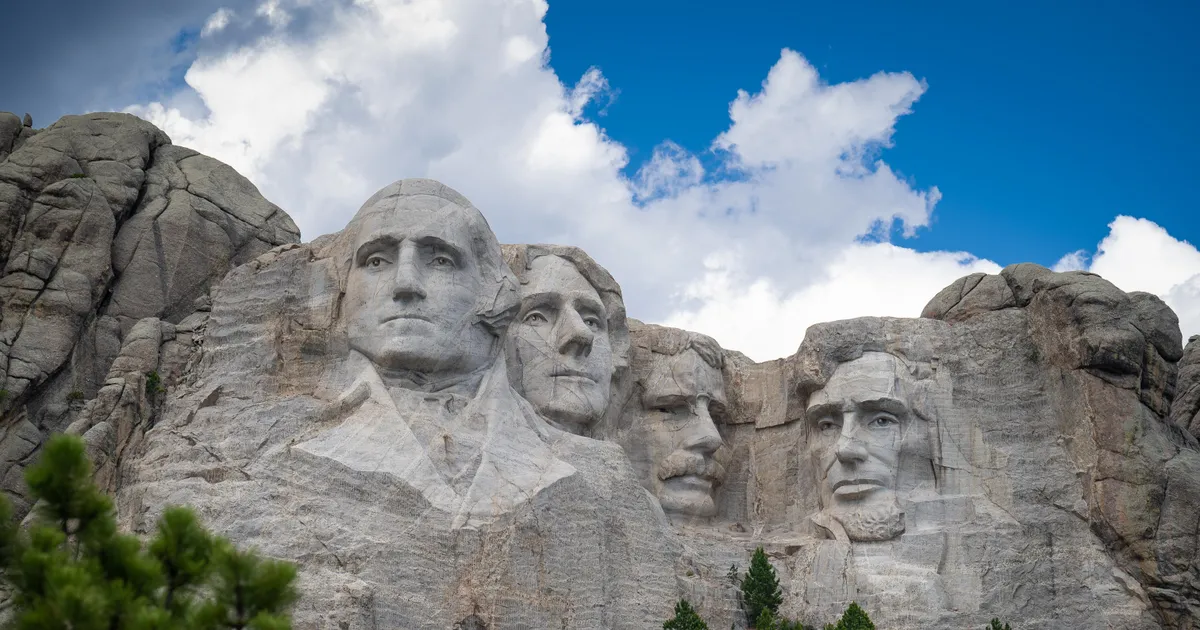 Photo of Mount Rushmore under a bright blue sky with puffy clouds floating over.