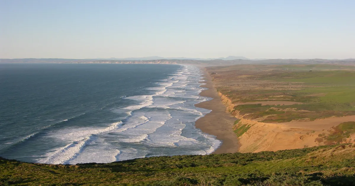 Waves wash in from the left onto a long, straight stretch of undeveloped, low-lying coastline.