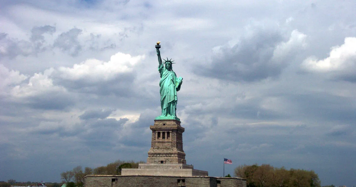 Patina green statue atop her pedestal on Liberty Island; grey cloudy sky in the distance